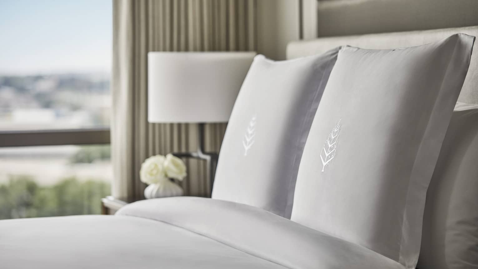Close-up of hotel bed, pillows with embroidered Four Seasons logo, nightstand with white roses