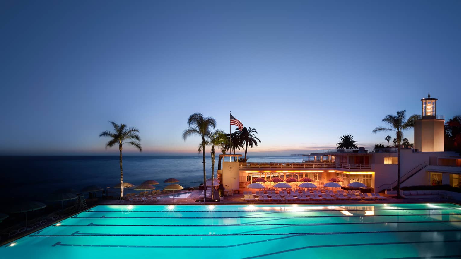 Illuminated Olympic-sized swimming lane pool, Cabana Club patio. palms and ocean at dusk