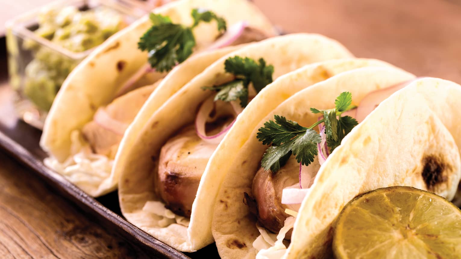 Row of three fish tacos, guacamole, lime on tray