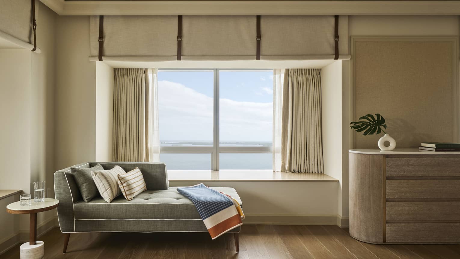 A chaise longue with blanket is positioned next to a picture window with view of the sea, in a hotel suite
