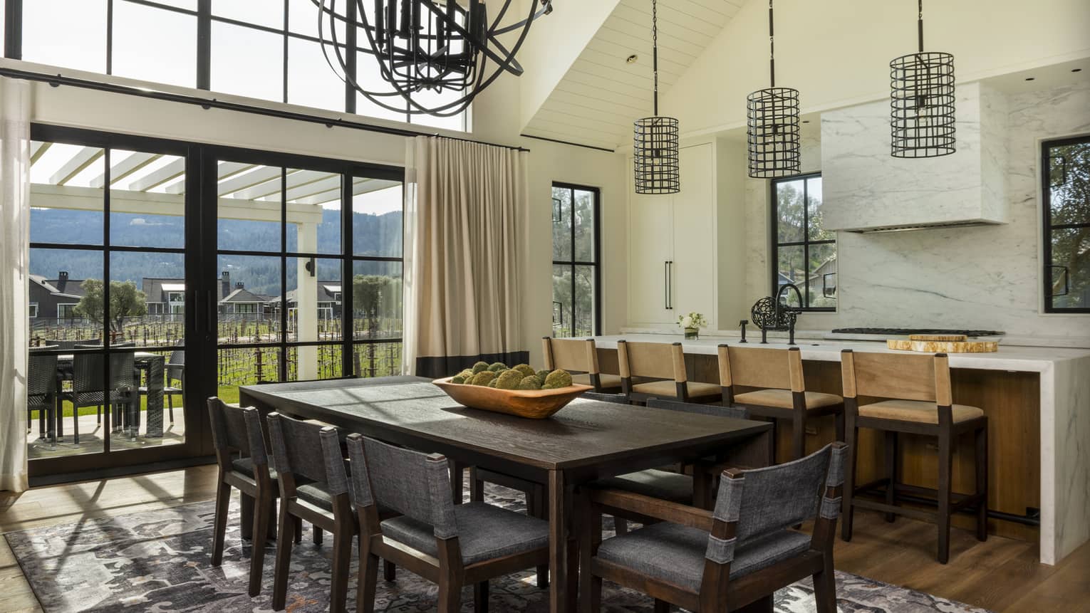 Dining and kitchen area with dark wood table, gray chairs, kitchen island with barstools, pendant lights, windows and sliding doors opening to outdoor patio 