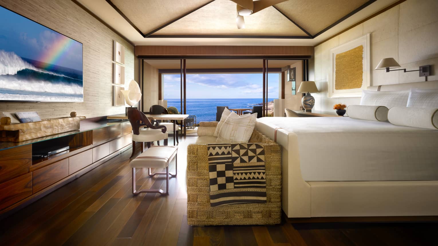 Oceanfront Room bed, small sofa and bench at foot, TV screen displaying ocean waves, rainbow