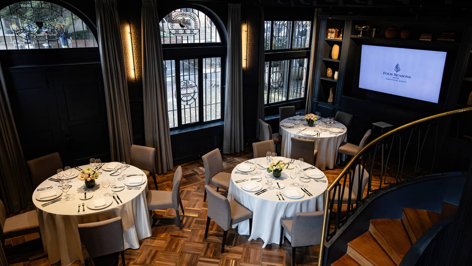 A spacious event room with round tables set for a meal, chairs, large arched windows, decorative wood flooring and a flat-screen TV