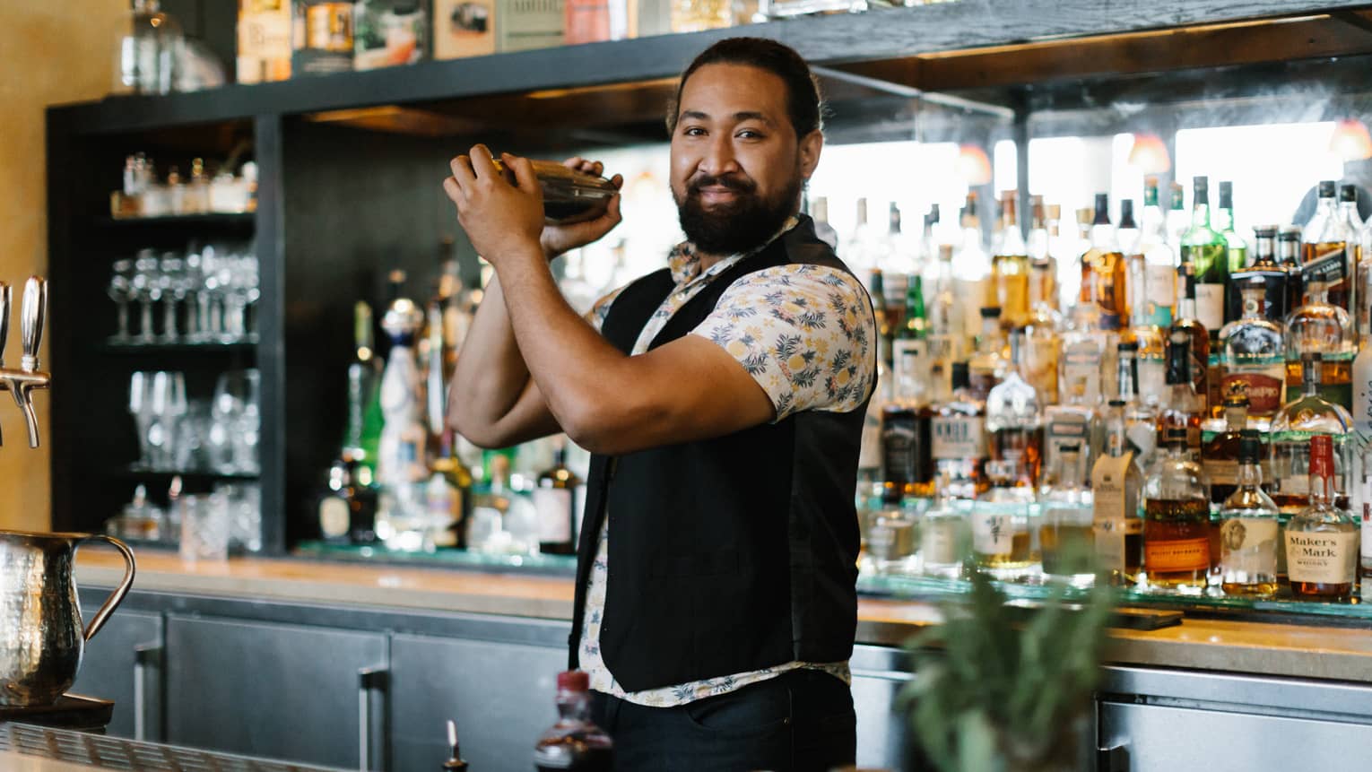 A bartender shaking a drink with a large assortment of bottles behind him.