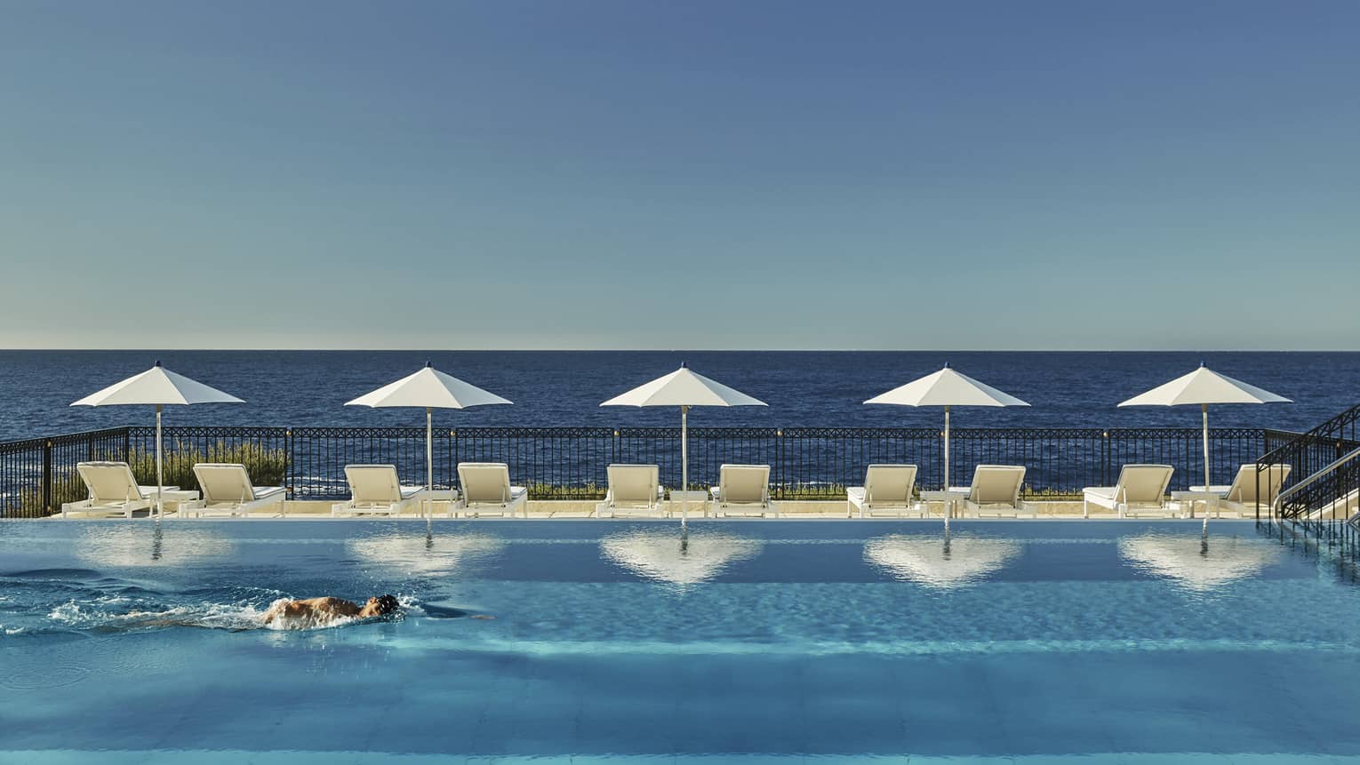 A man swims in outdoor lap pool, lined with lounge chairs and white umbrellas, looking out to sea
