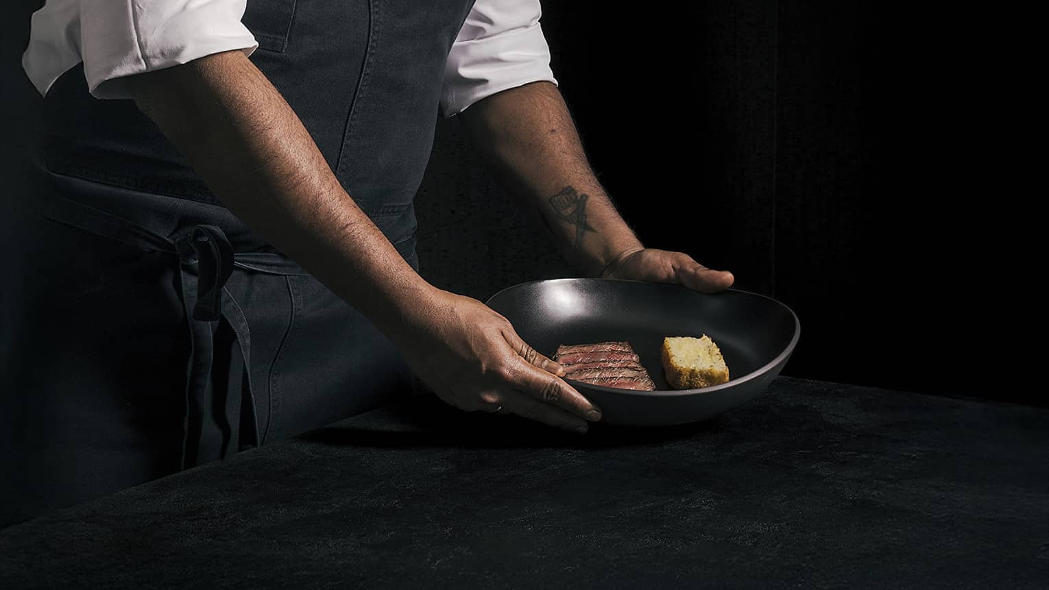 Chef wearing a white, long-sleeve chef's jacket and a dark denim apron places a black plate of food onto a black table