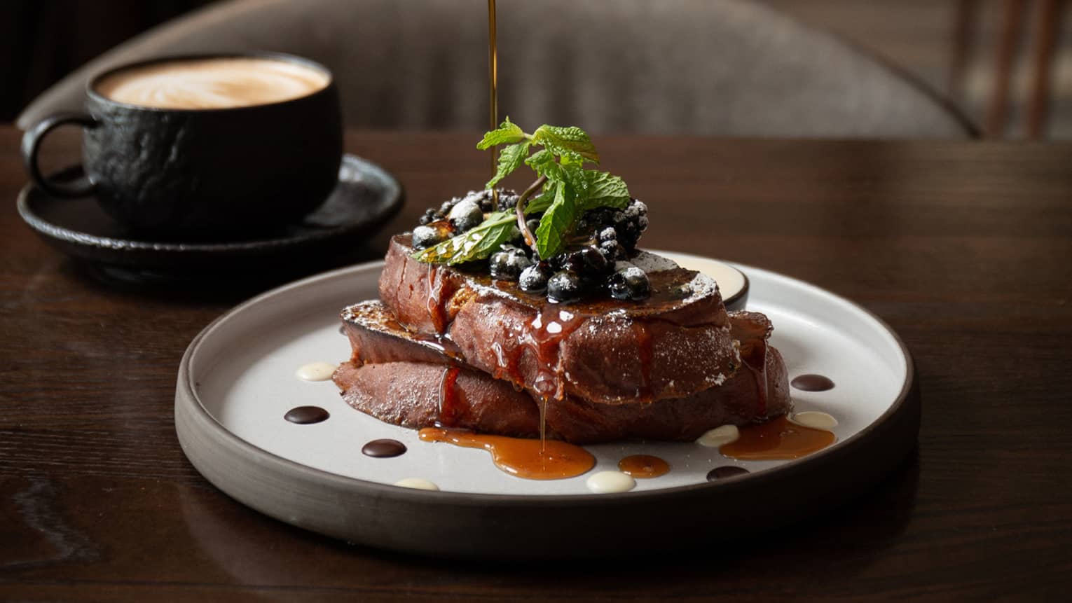 A plate of French toast sits next to a cup and saucer on a dark brown table. Syrup is being poured over the French toast.