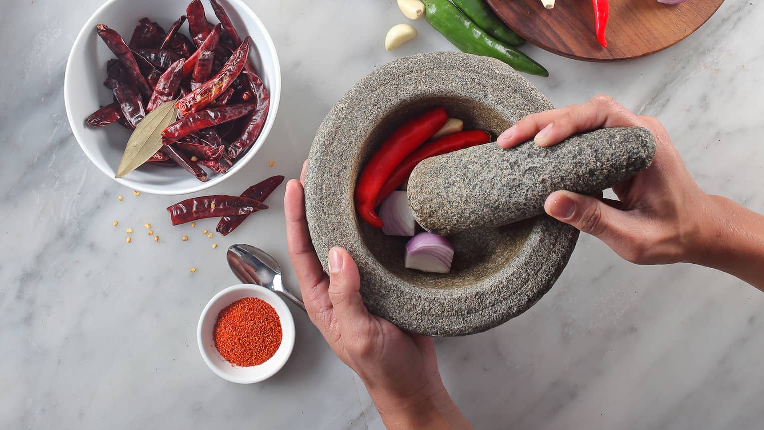 White marble counter with spices, woman crushing red onions, hot peppers with mortar and pestle