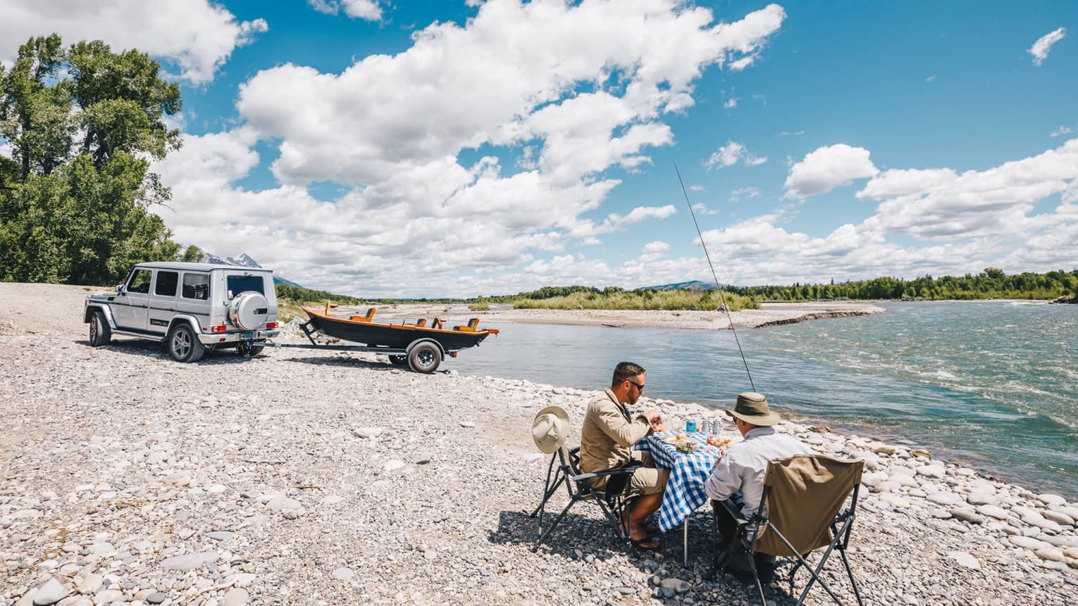 Two fishermen sit at a small table for a lakeside picnic, their boat hitched to their SUV in the background