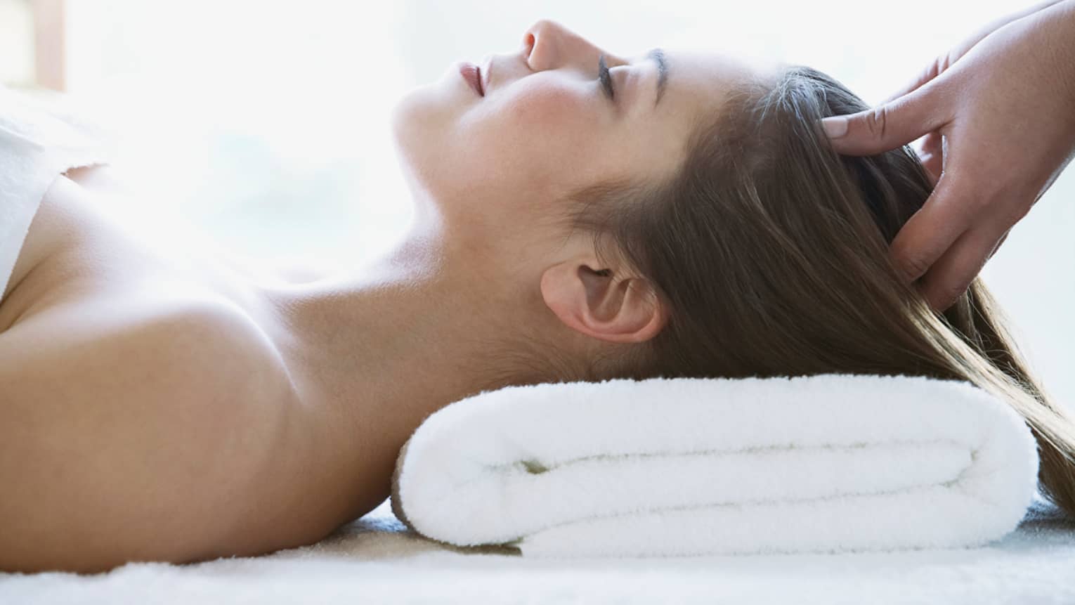 Woman lays on back with head on folded white towel, closes eyes as hands massage her scalp