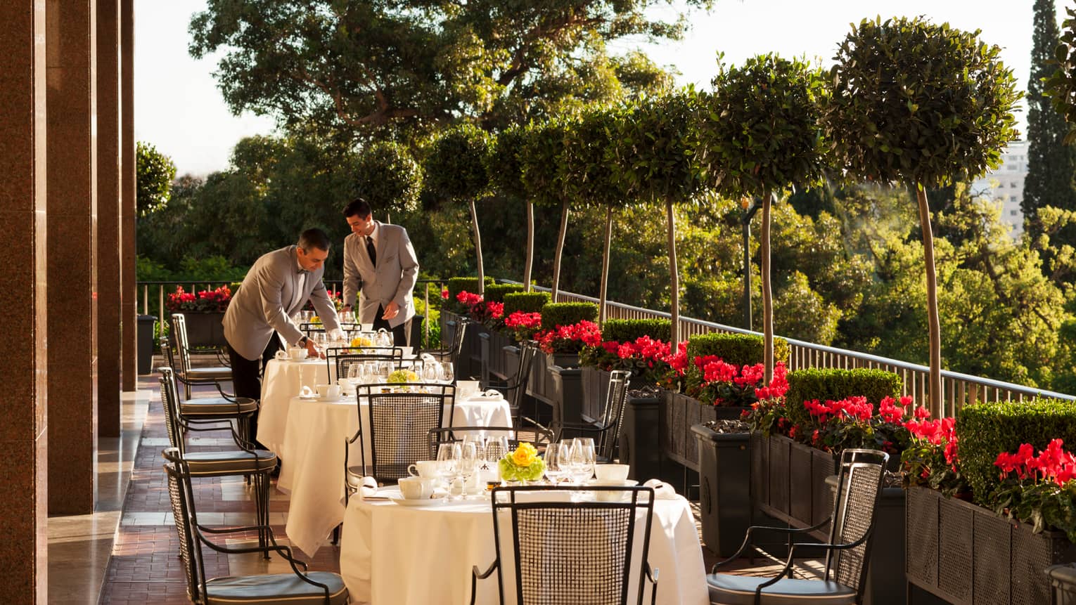 Two servers in grey jackets with black ties set outdoor dining tables with white tablecloths, dishes, red flowers along side