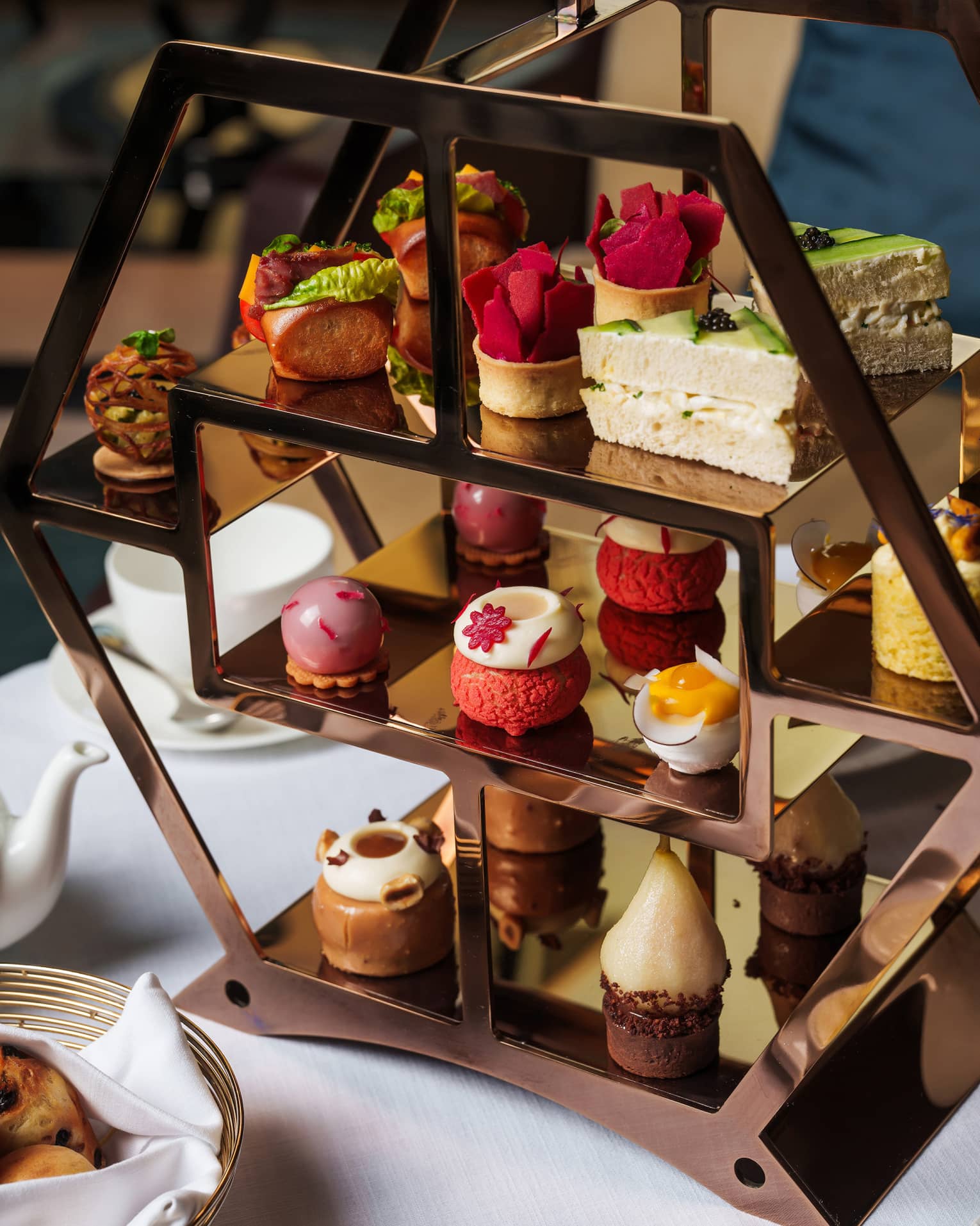 Bronze hexagonal three-tiered tray filled with small pastries and set on a table next to small containers of jam and a silver bread basket