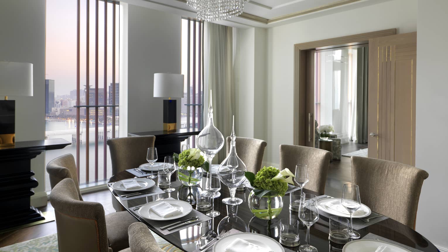 Elegant dining room with a glass-top table, beige chairs, crystal chandelier and floor-to-ceiling windows offering a city view at sunset