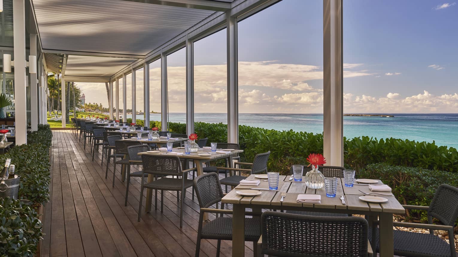 An outdoor eating area with square wood tables next to plants and the ocean.