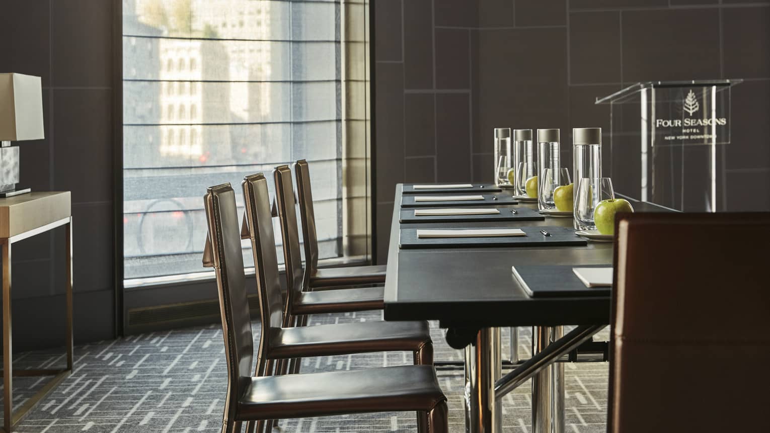 Dark table with chairs, stationery, bottles of water, green apples in light-filled meeting room with podium