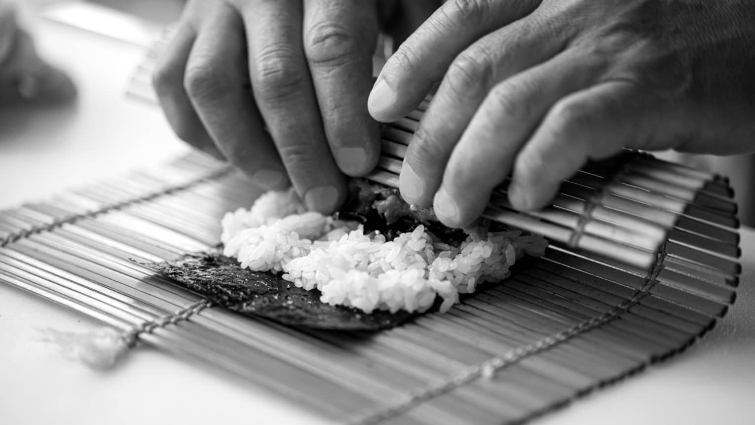 A black and white close up image of a handing rolling sushi in a bamboo mat 