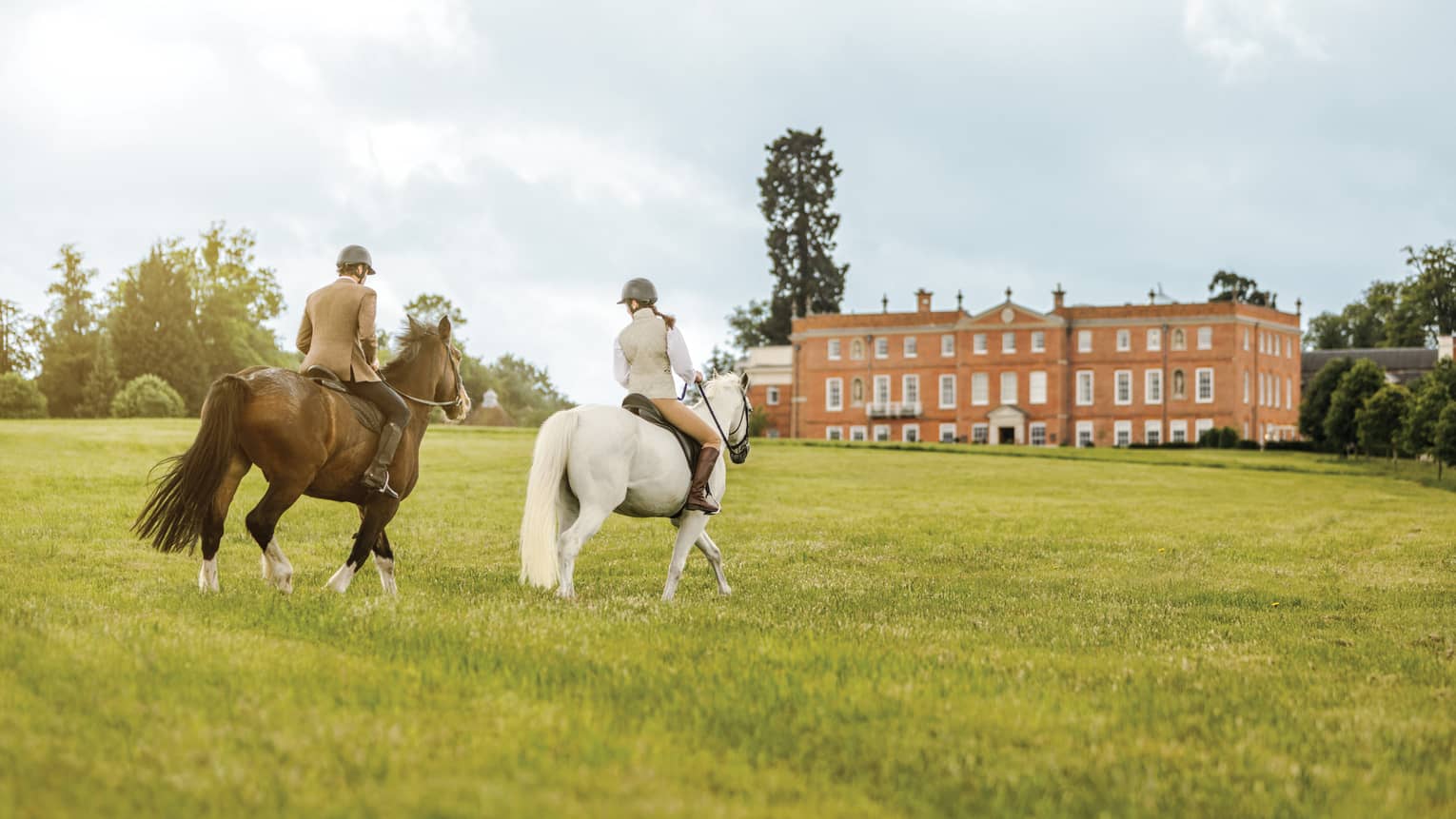 Couple rides horses across green English field to Four Seasons Hotel Hampshire building