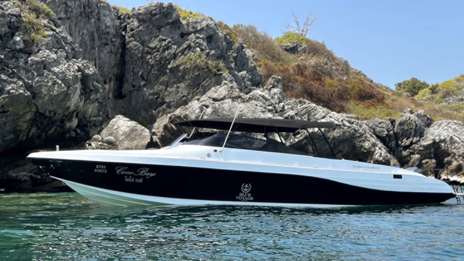 Side view of an elegant speedboat with a canvas roof floating in the ocean alongside a rocky cliff and scrub-grass