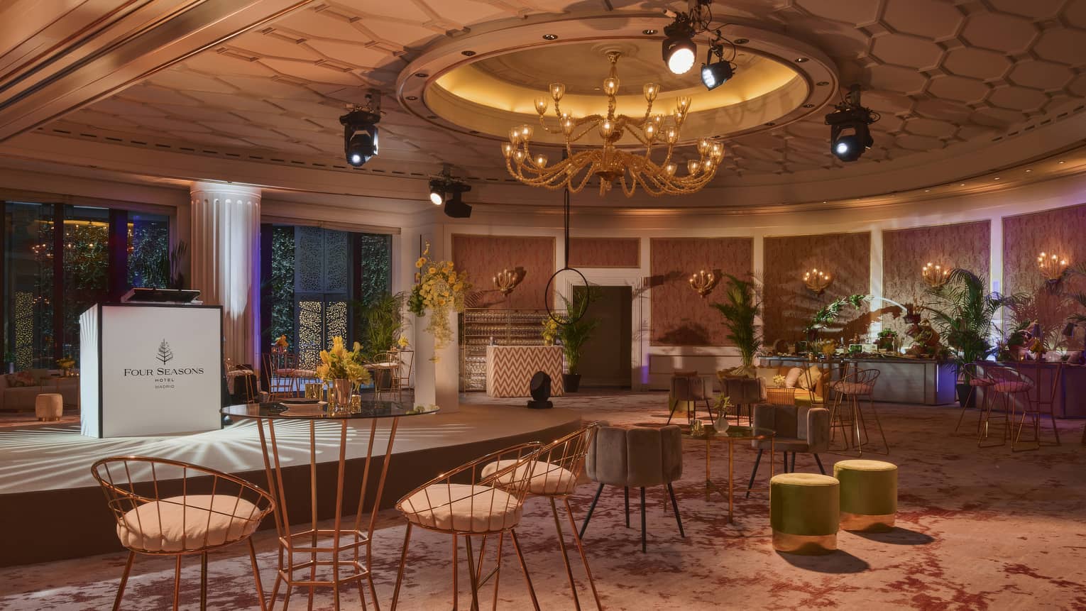 A large, open meeting space warmly lit with overhead and wall sconce lighting also features large windows, greenery and floral arrangements, a small stage with a podium, and table and chair groupings.