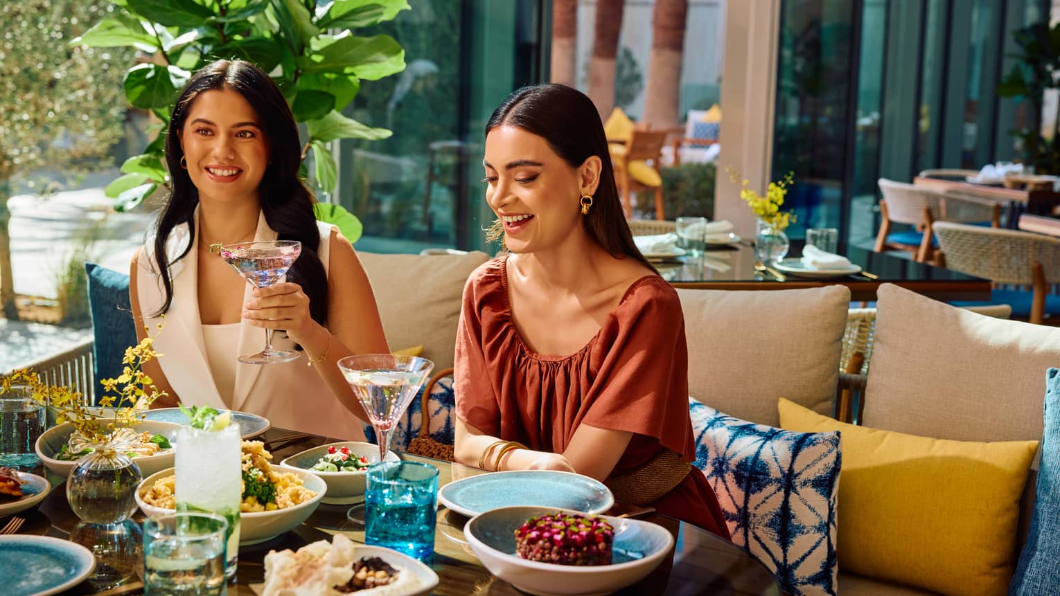 Two guests sit on a sofa filled with yellow and blue throw pillows set next to a table filled with food on an outdoor restaurant patio