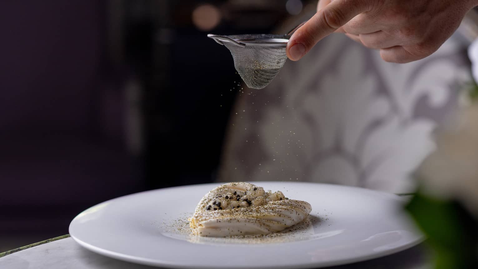 A person sifts a topping onto a food item on a round white plate.