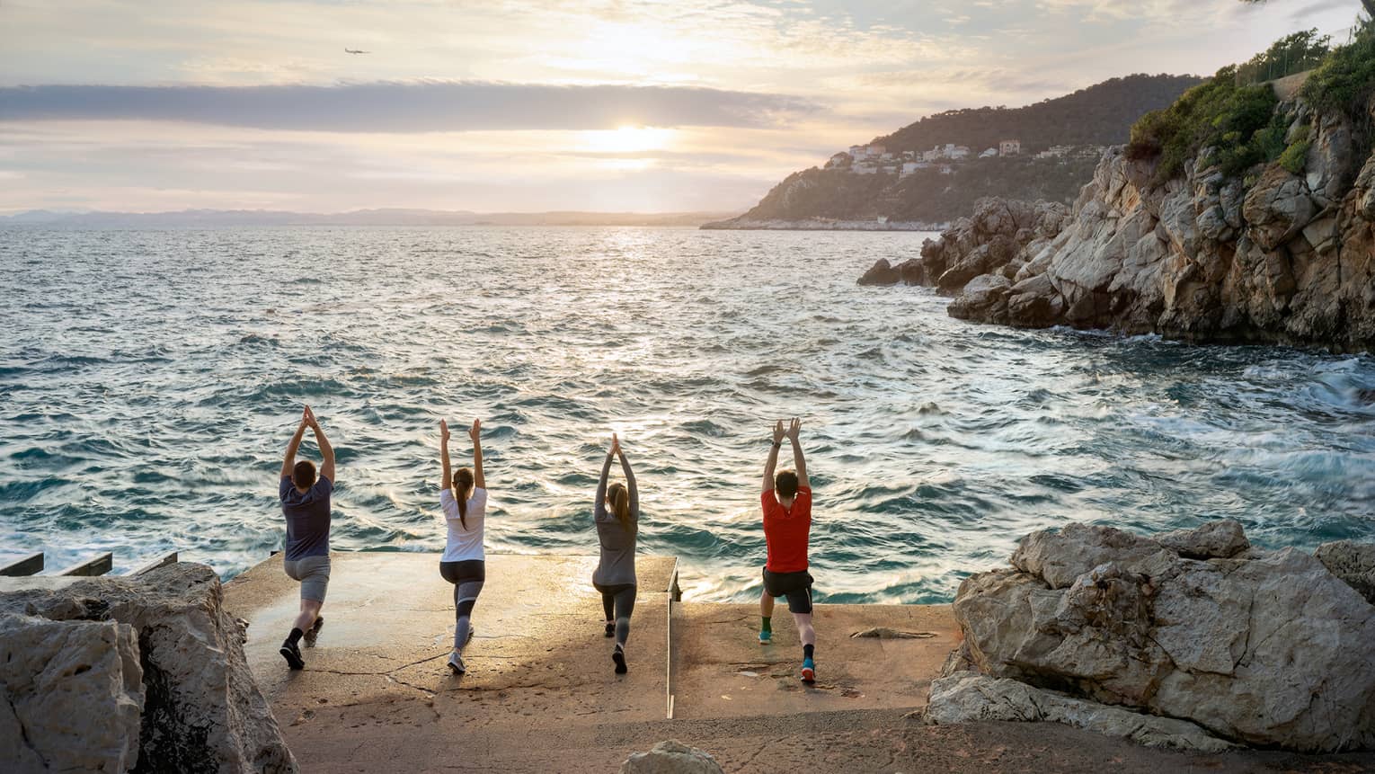 Rear view of four people doing yoga on sandy shore next to sea, rocky cliffs on right