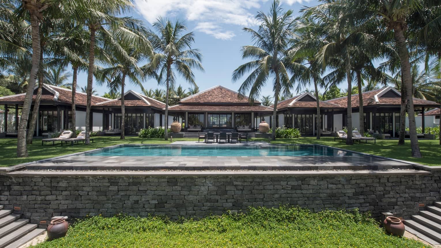 Luxurious villa with a large swimming pool, surrounded by palm trees and greenery, featuring modern architecture with tiled roofs and outdoor seating areas.