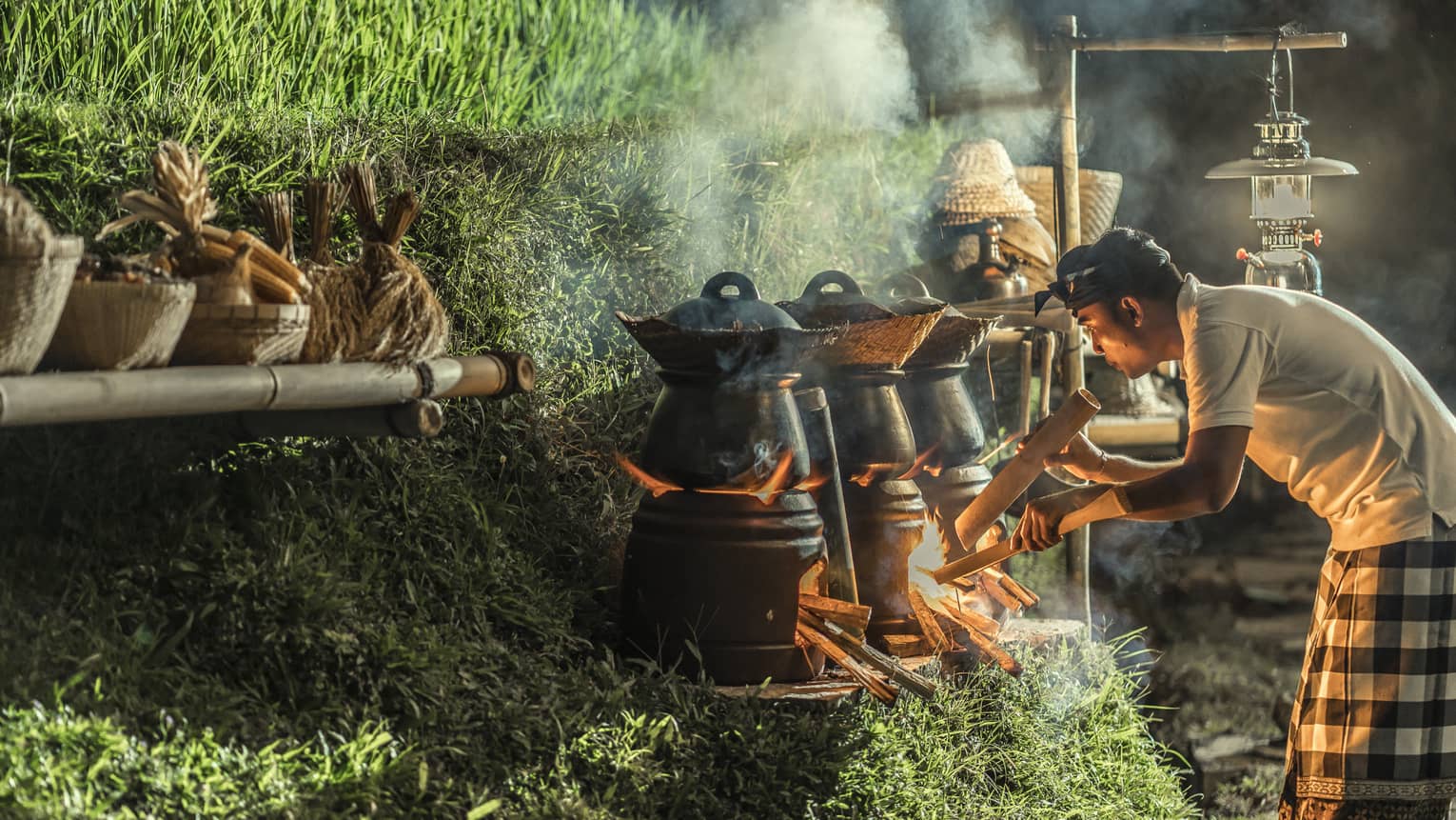 A chef stokes the fire beneath a row of outdoor cooking pots set on a grassy hill, the smoke illuminated by the flames.
