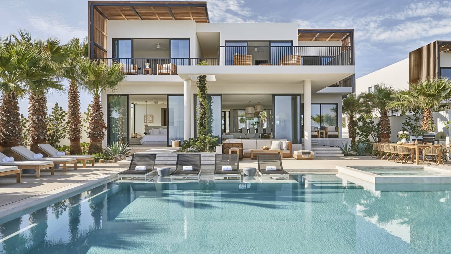 Modern luxury villa with a spacious pool area, surrounded by palm trees, featuring outdoor lounge chairs, a dining area, and floor-to-ceiling windows.