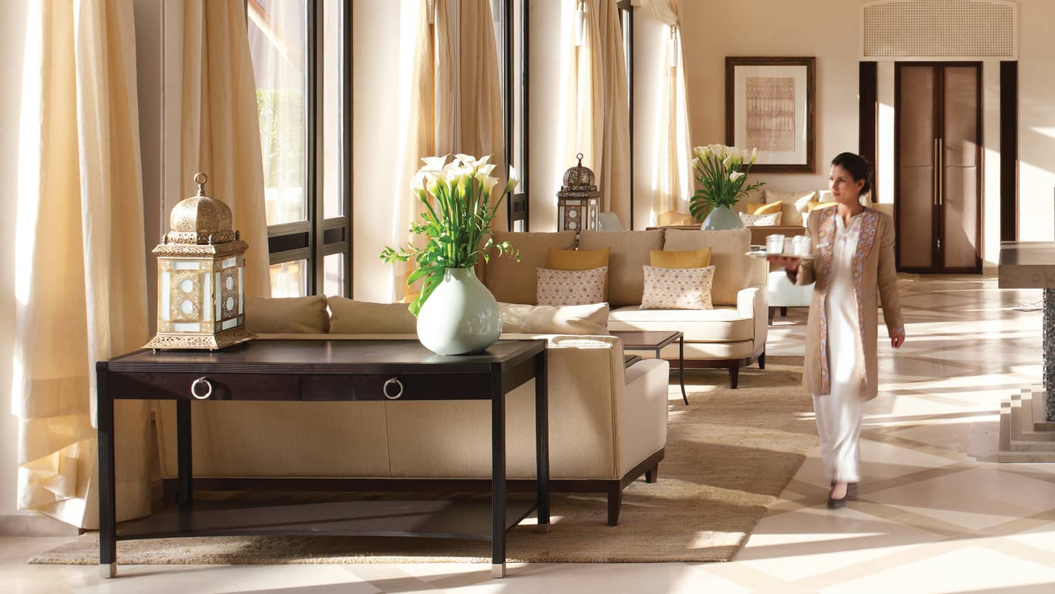 Server with tray walks through sunny lounge past white sofas 