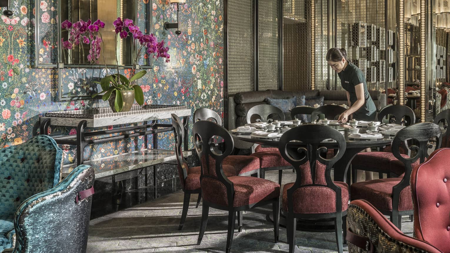 Hotel staff sets table with elegant red velvet chairs under tall mirror, floral wallpaper 
