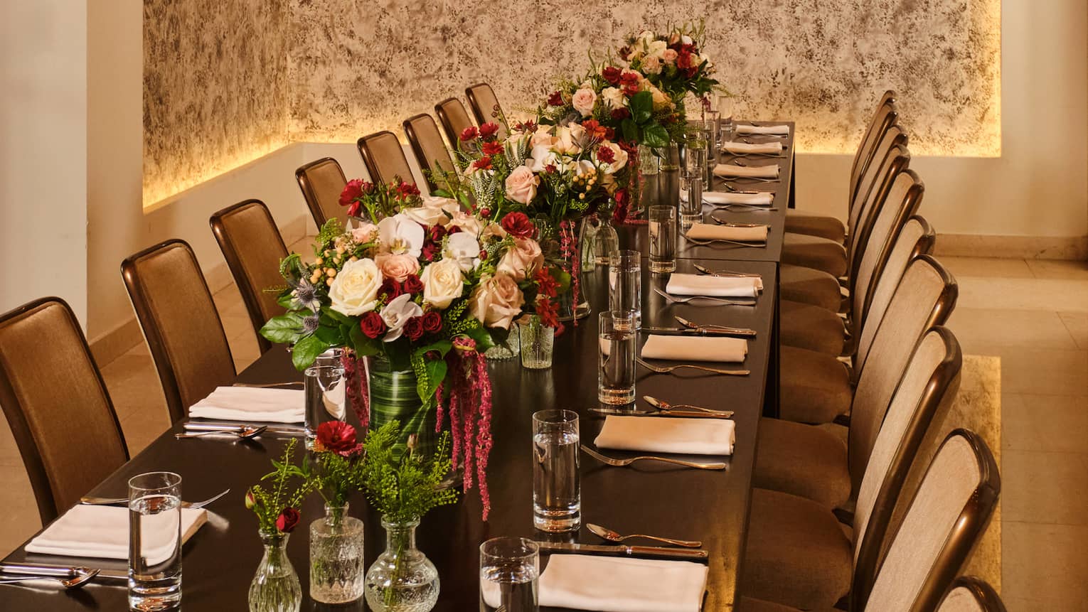 Long rectangular dining table set in a private dining room with red-and-light-pink floral arrangements in the centre and string lights overhead