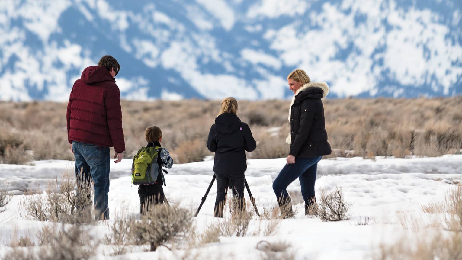 Two children stand between man and woman wearing winter coats looking at snowy mountain view 