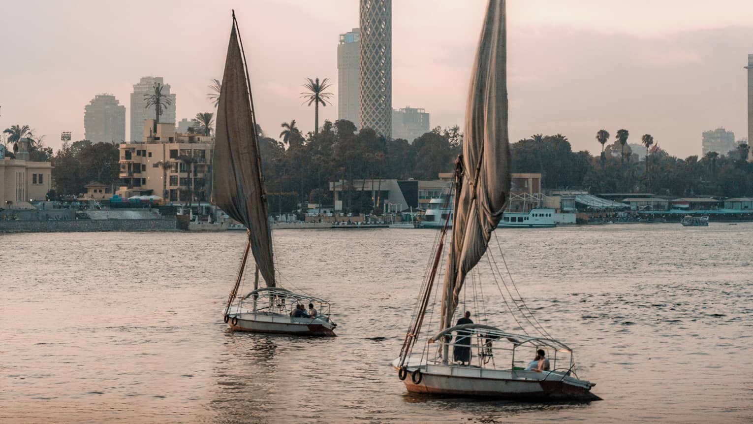 Two felucca boats drift on the water in the evening light, a busy shoreline and Cairo Tower in the distance. 