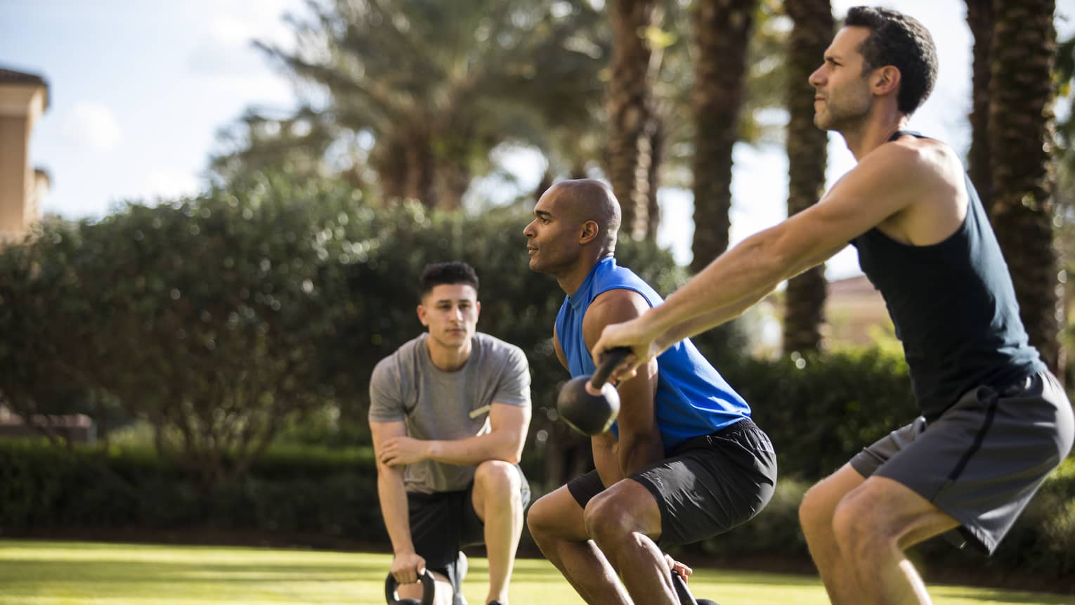 Four Seasons trainer watches as two men squat, lift kettlebell weights on lawn