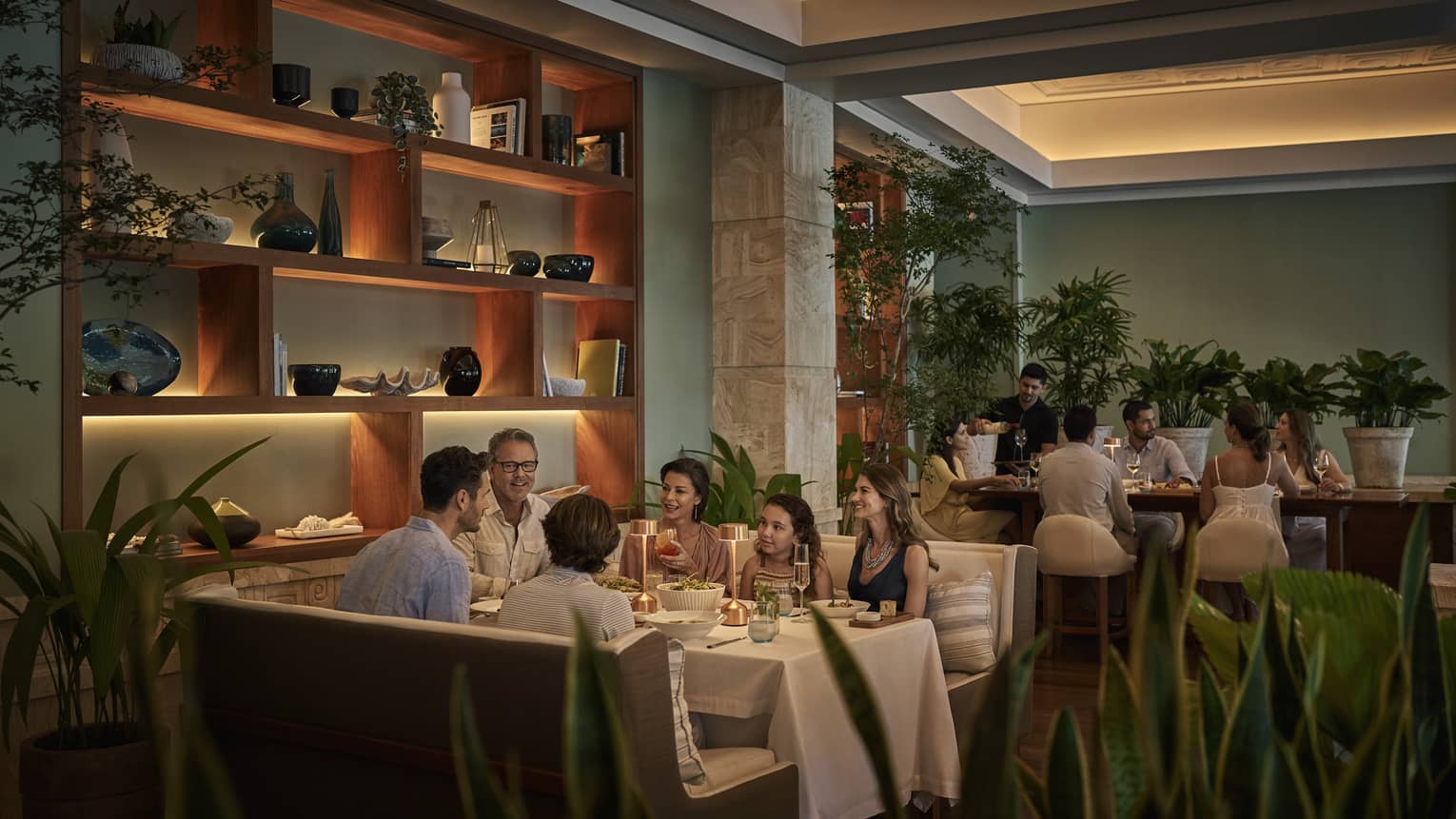 A family of six sits at a dinner table at Pesce, flanked by various green plants and a wall fitted with wooden shelves filled with various ceramics