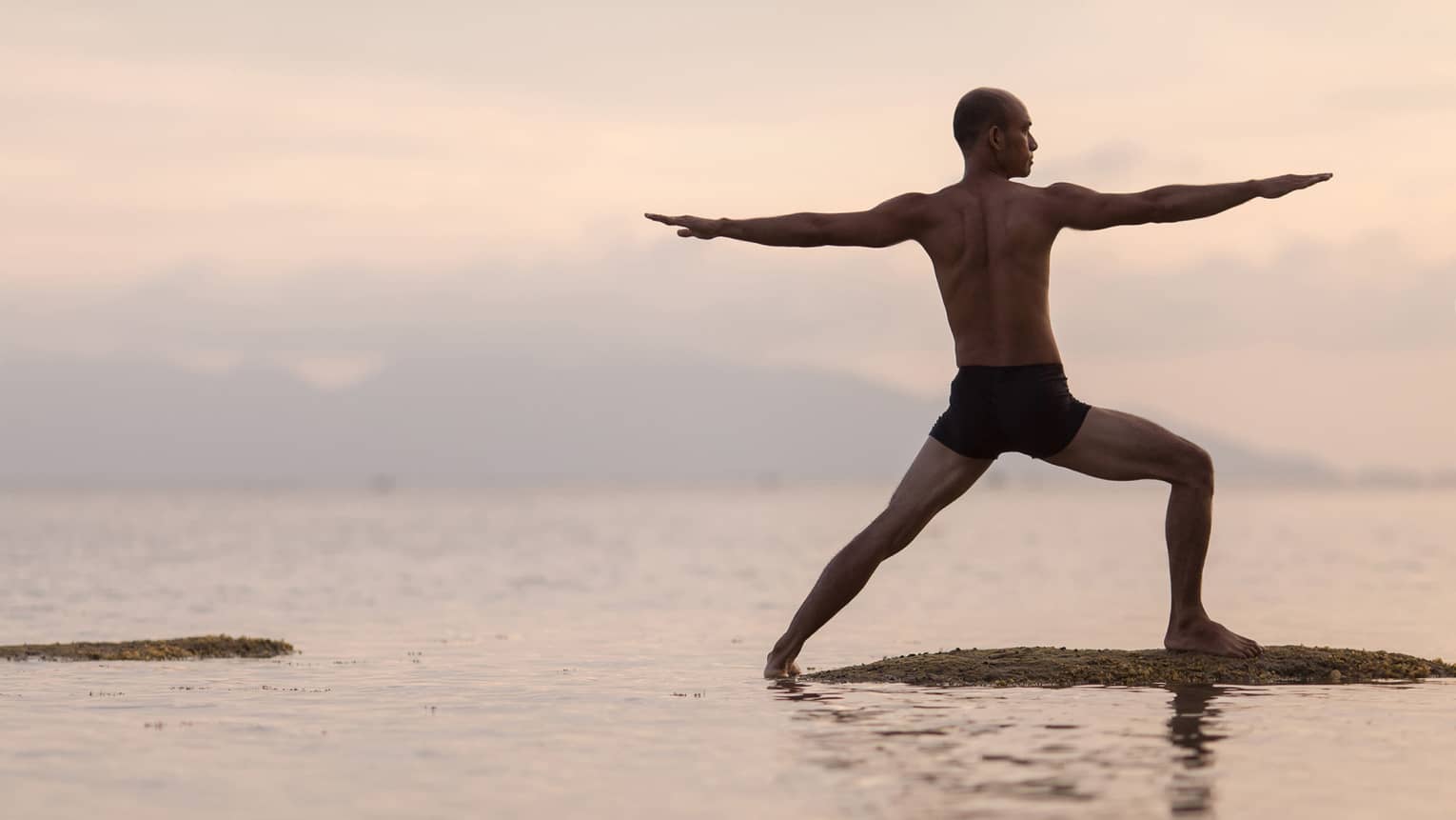 Silhouette of man in swimsuit doing yoga pose on sandbar in ocean at sunset 