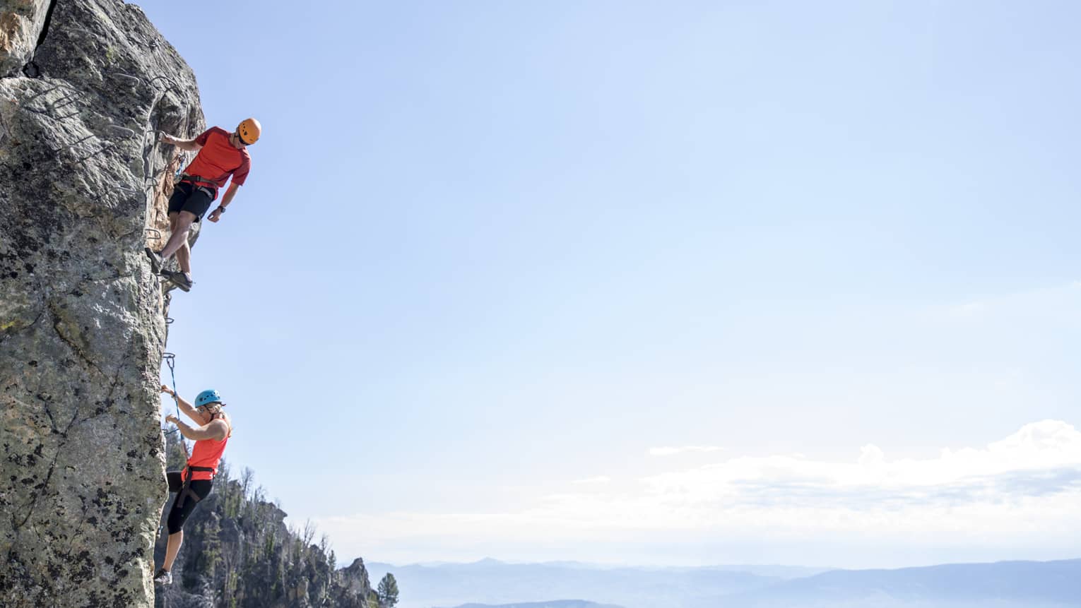 Man and woman climb steep cliff face with view of mountains in far distance 