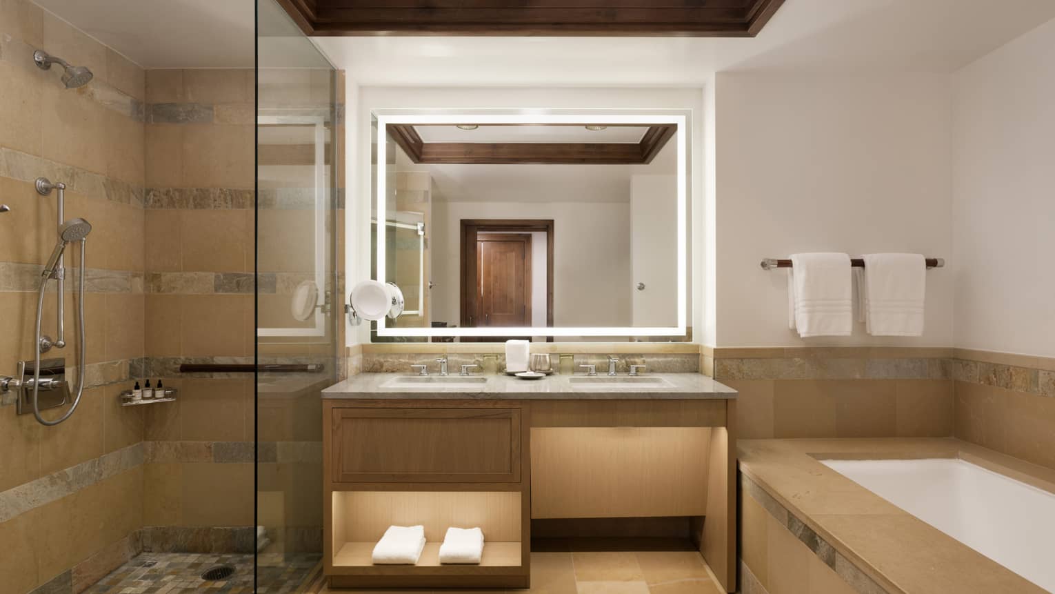 Spacious bathroom with a double vanity, glass-enclosed shower and a beige tiled bathtub, illuminated by a backlit mirror