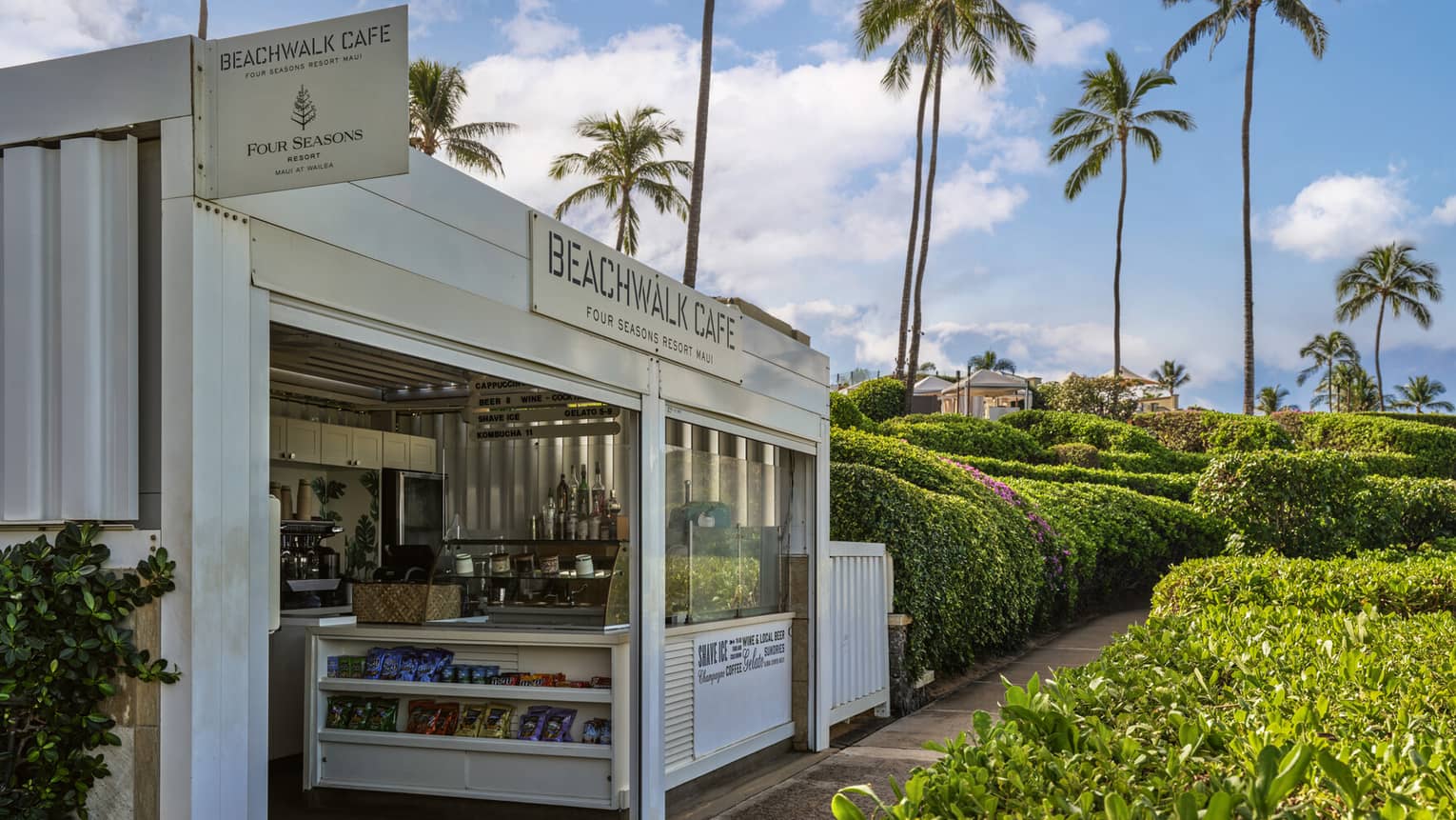 Exterior of a walk-up cafe and shop by rows of lush greenery and a paved pathway