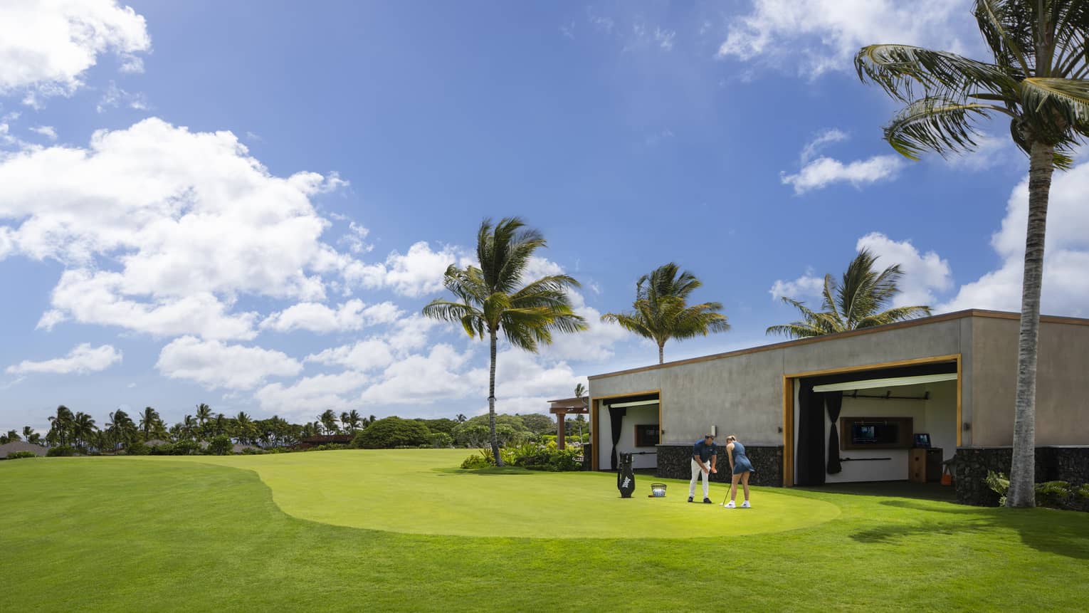A woman receives an individual golf lesson on a golf course outside a building with palm trees.