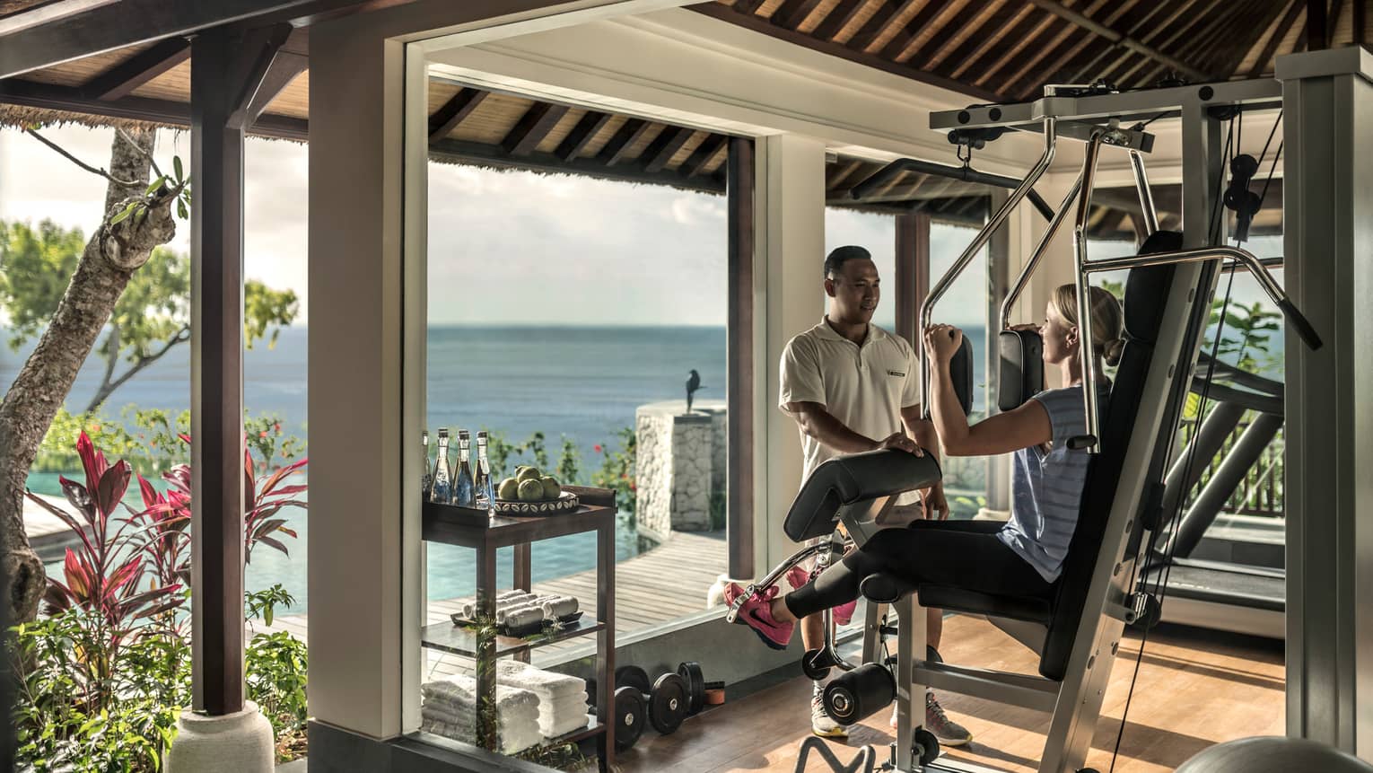 Imperial Villa private gym, trainer assists women on weights machine by window with ocean view