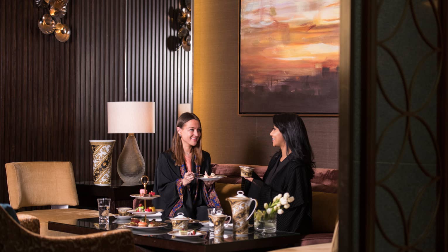 Two women in thin black robes sit on sofa, enjoy high tea at table
