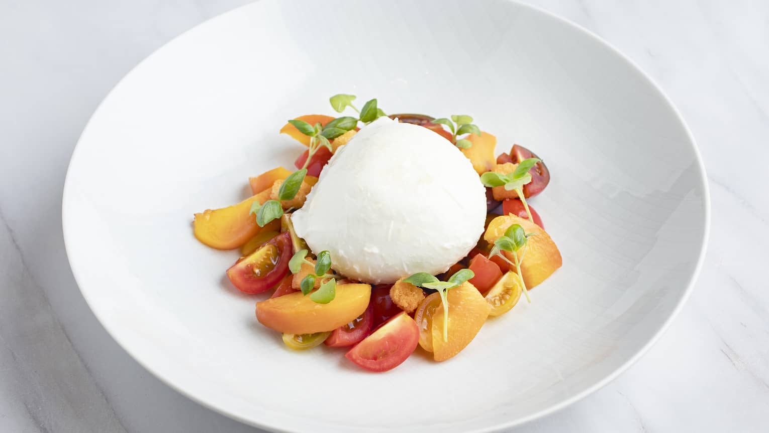 Ball of fresh mozzerlla, sliced cherry tomatoes, roasted peach slices, basil pesto, croutons and sprigs of fresh basil plated neatly in a white wide-rimmed bowl