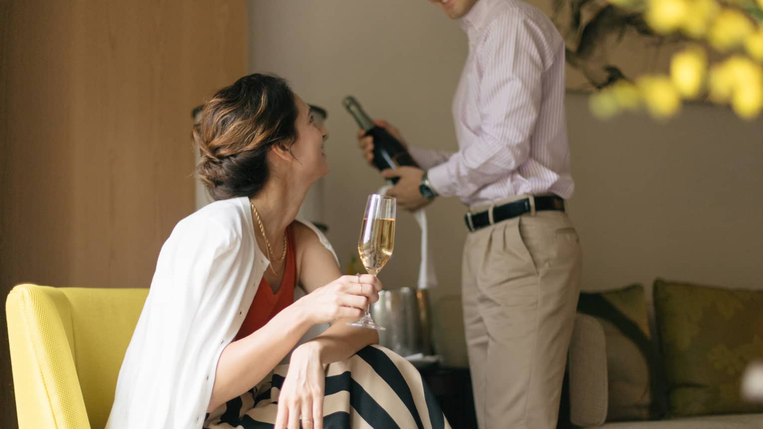 Person smiling and holding a glass of champagne while another person opens a bottle