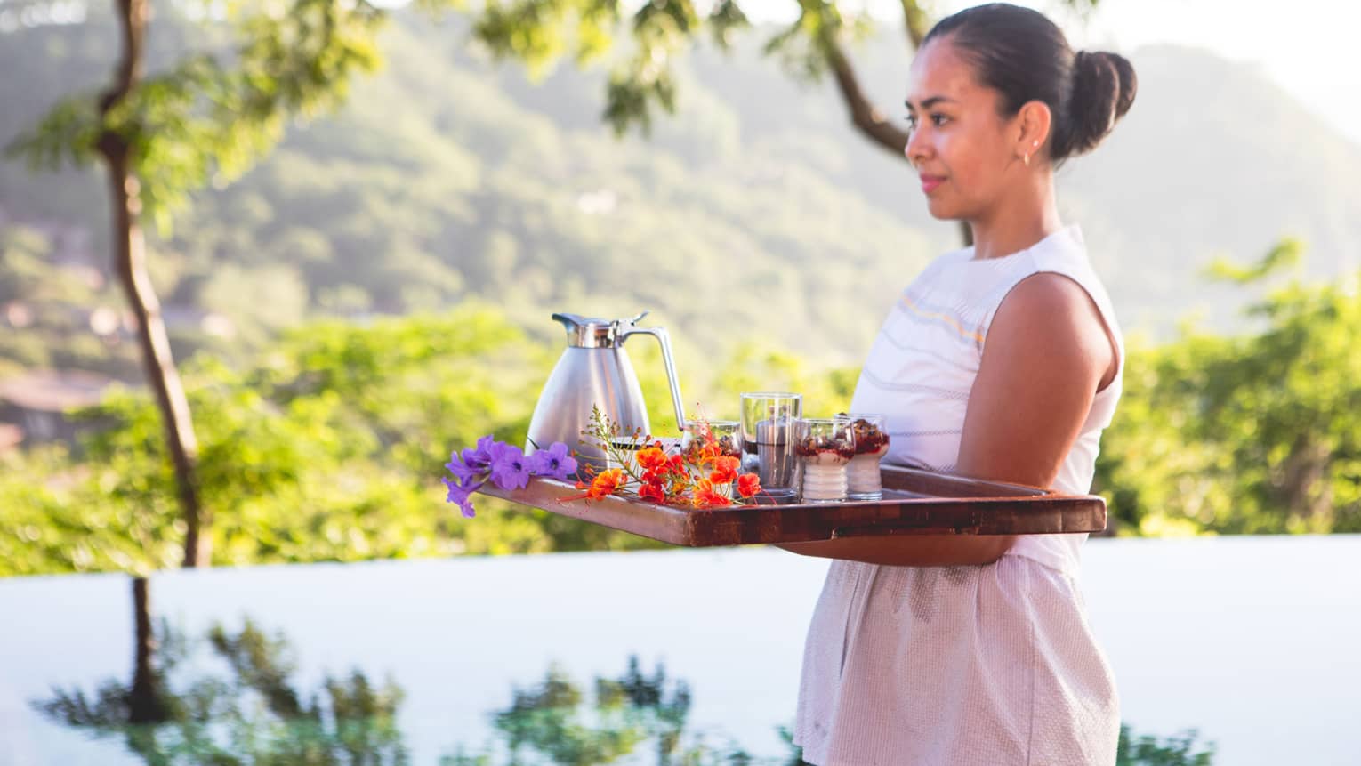 Woman carries breakfast tray with tropical flowers past outdoor pool