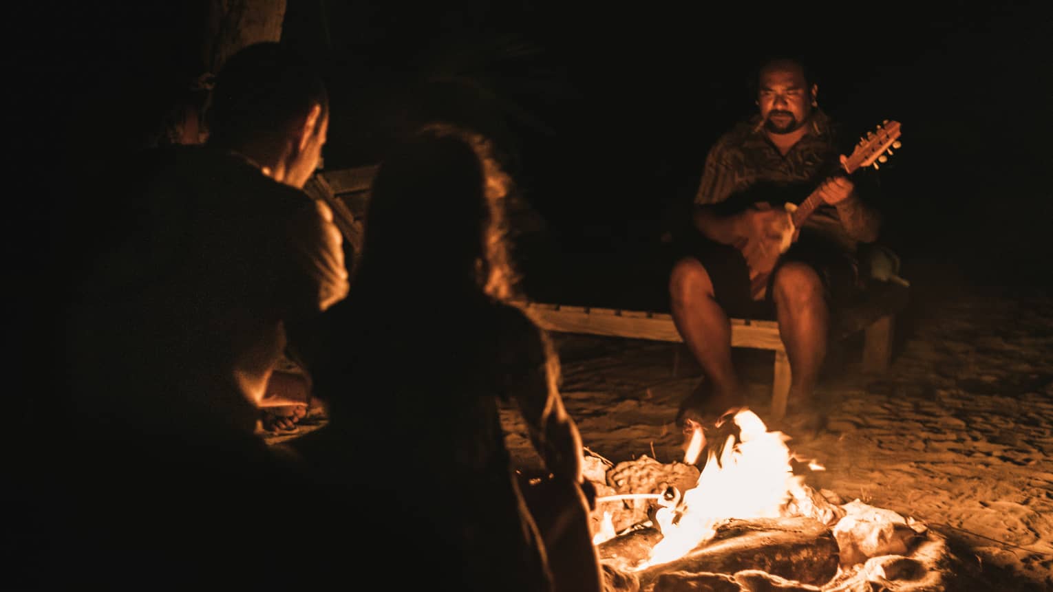 Group of people sitting around a campfire at night, with one person playing a guitar in the background