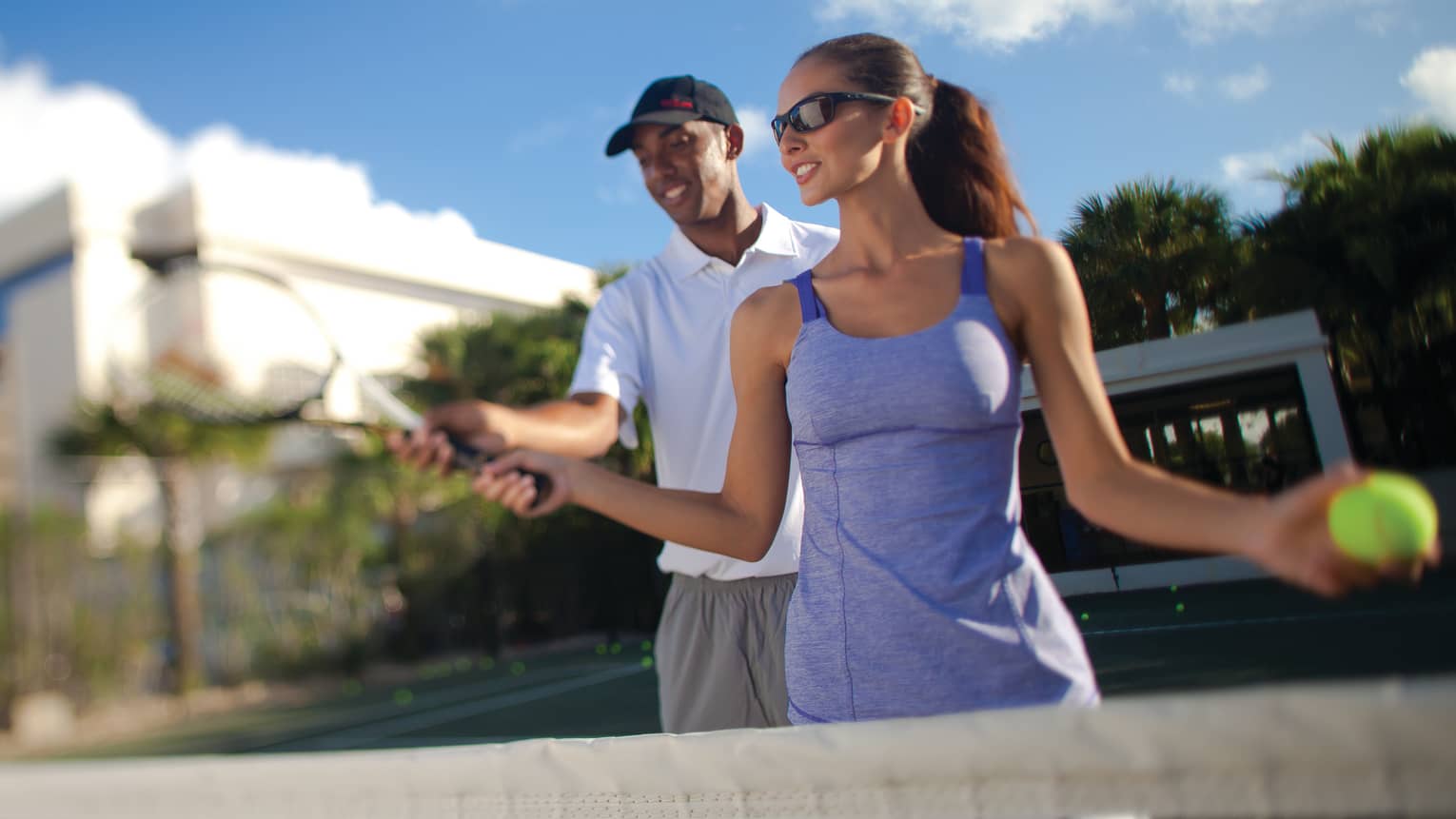 Man helps woman with tennis racket on court under blue sky