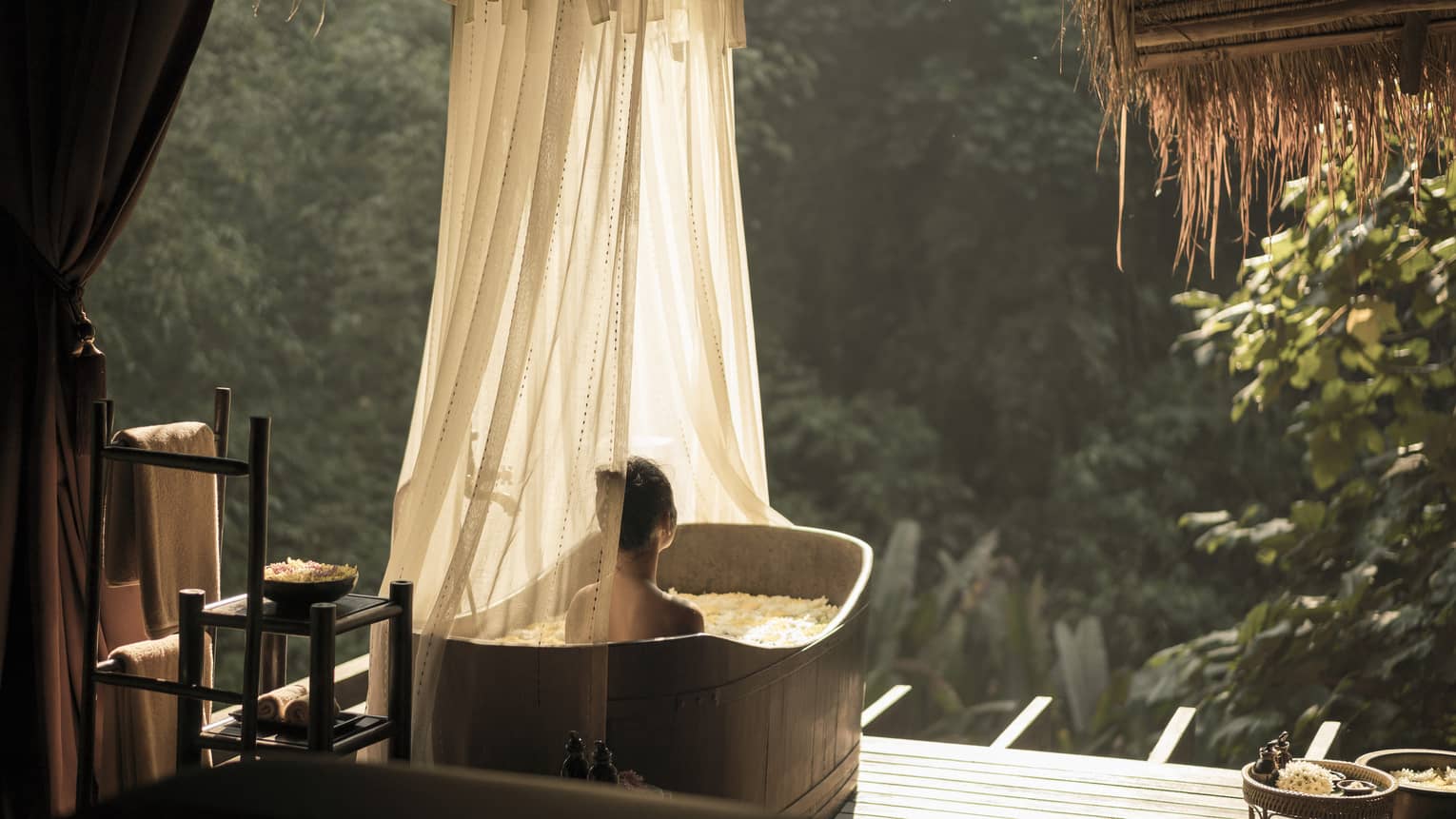 A person relaxes in an outdoor bathtub with sheer curtains, surrounded by lush greenery and sunlight. Towels and bath items are neatly arranged nearby, creating a serene spa atmosphere.
