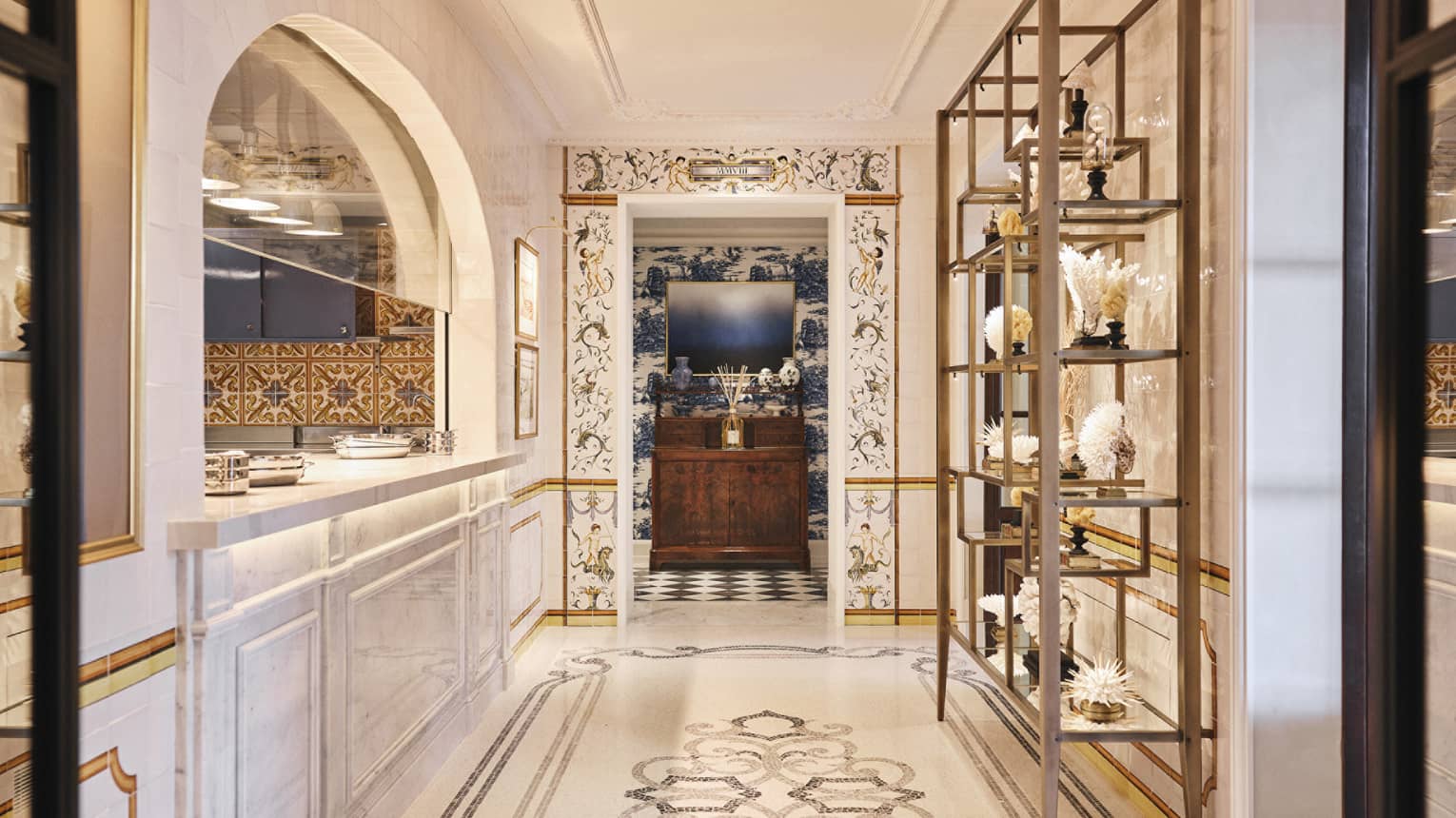 A long, bright hallway with a floor of decorative tile work. A an arched opening with a counter is along one wall and display shelves are along another wall.